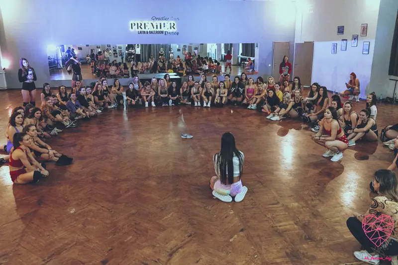 Group twerk class drilling pelvic tilts in front of a studio mirror with hands on hips for tactile feedback — the standard isolation drill for unlocking hip motion